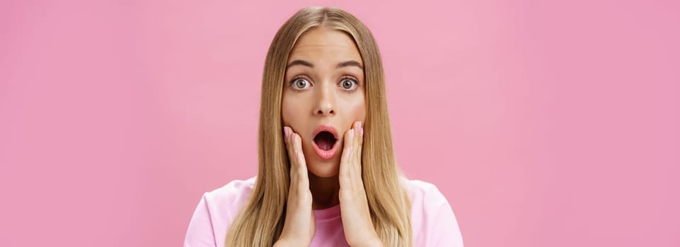 Close-up shot of shocked young female student with tanned skin and fair hair dropping jaw gasping from amazement touching cheeks surprised reacting to shocking stunning news over pink background. Copy space