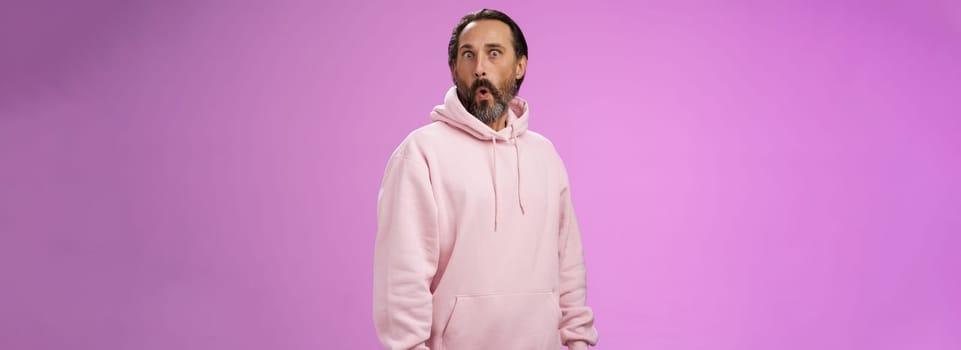 Impressed speechless shocked adult bearded man grey hair in cool pink hoodie folding lips wow popping eyes camera surprised astonished intrigued, standing curious amazed purple background.