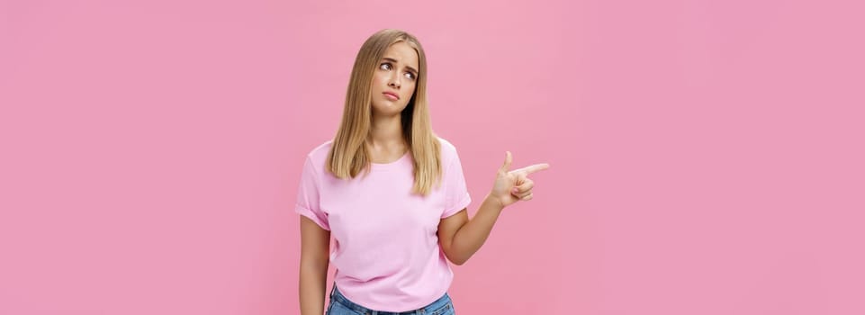 Envy upset cute young european woman with tanned skin and fair hait tilting head lifting eyebrows in sad silly look pointing, gazing left with regret and disappointment posing against pink background.