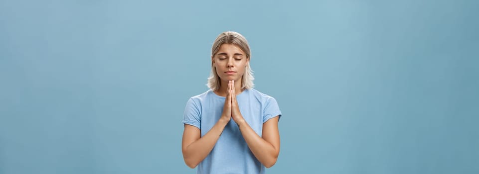 Lifestyle. Good-looking kind and faithful european girl with blond hair smiling cute and tender holding hands in pray while making wish hopefully believing god hearing her prayers with closed eyes over blue wall.
