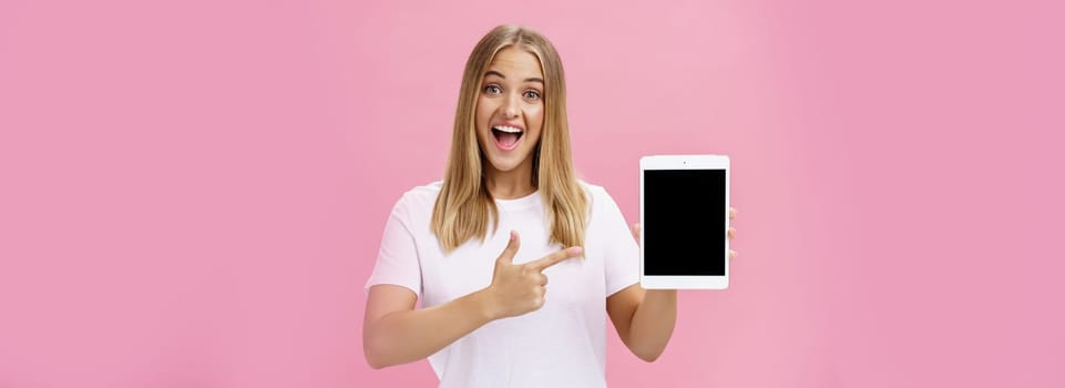 Woman giving recommendation buy digital tablet pointing at device screen with index finger smiling enthusiastic and pleased telling own opinion about gadget perfomance posing delighted over pink wall. Lifestyle.