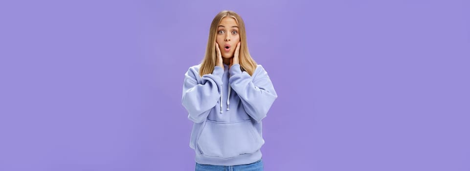 Indoor shot of amused and thrilled surprised good-looking young woman in stylish hoodie folding lips in wow sound popping eyes at camera shocked and excited learning amazing news over purple wall. Lifestyle.