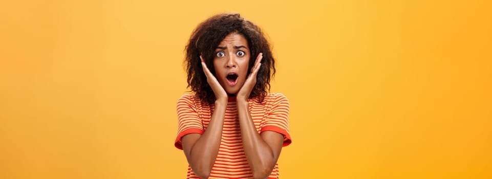 Waist-up shot of shocked concerned panicking african-american woman with afro hairstyle in trendy t-shirt gasping holding palms on face from surprise posing troubled against orange wall. Lifestyle.