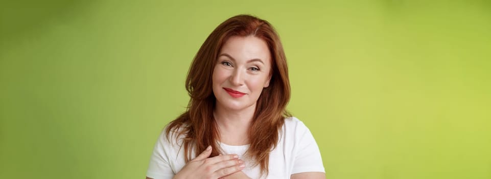 Lovely pleased tender redhead middle-aged ginger woman. press hands heart feel touched grateful appreciate sincere heartwarming moment admire thankfully grin look camera glad green background.