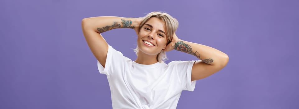 Lifestyle. Close-up shot of good-looking stylish and relaxed blond woman with tattoos on arms holding hands behind head smiling with carefree pleased look enjoying weekends over purple background.