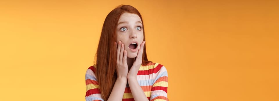 Shocked amazed young drama girl redhead blue eyes gasping empathy touch cheeks shocked drop jaw worry turn left surprised astonished standing nervously orange background. Emotions concept