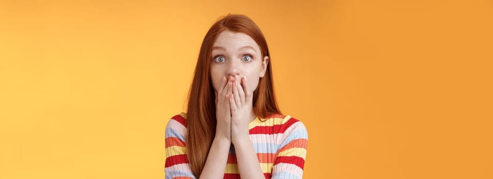 Shocked speechless impressed sensitive redhead european girl reacting stunning rumor gossiping find out secret gasping cover mouth palm stare camera astonished surprised, orange background.