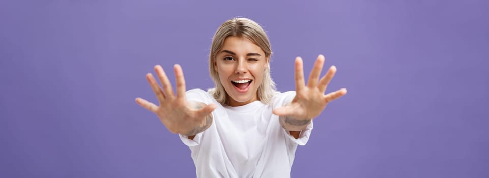 Reaching to you. Portrait of flirty good-looking and confident stylish woman with tattoos on arms winking sticking out tongue playfully and smiling pulling hands towards camera over purple background. Emotions and body language concept