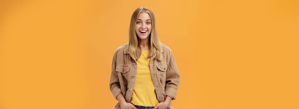 Charismatic tanned woman in corduroy jacket and yellow t-shirt ready for chilly autumn walk with friends smiling joyfully gazing entertained at camera holding hand in pockets casually over orange wall. Lifestyle.