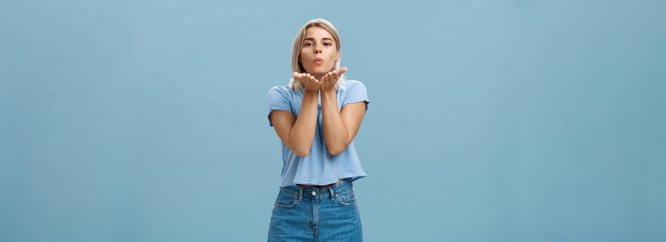 Lifestyle. Studio shot of attractive and romantic european blonde girlfriend in denim shorts folding lips sending wind kiss with palms near mouth gazing tender and cute at camera over blue background.