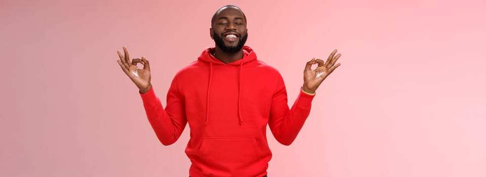 Happy african american bearded man in red hoodie meditating found peace nirvana smiling delighted close eyes relaxed relieved standing lotus mudra pose search zen, practice yoga, pink background.