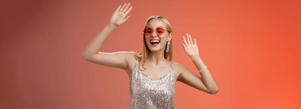 Happy amused carefree blond woman go wild dance-floor dancing having fun yelling yeah closed eyes waving hands moving rhythm music joyfully party in silver stylish dress sunglasses, red background.