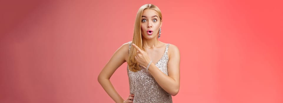 Wondered amused attractive blond woman talking party widen eyes say wow pointing questioned right direction see famous person standing surprised impressed gaze camera, red background.