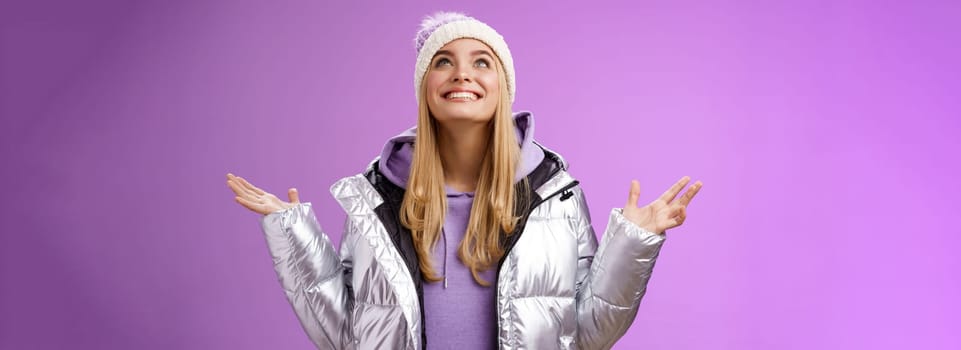 Thankful happy cute attractive blond young 25s woman in winter hat silver trendy jacket raise hands look up grateful god dream come true smiling delighted fulfilled wish, purple background.