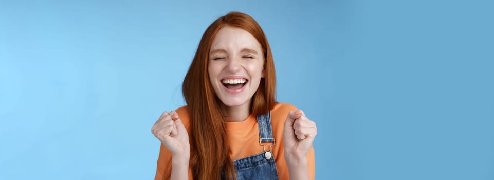 Lifestyle. Sincere happy rejocing ginger girl close eyes smiling broadly say yes waving clenched fists joyfully celebrate enterting university dream come true winning prize triumphing cheerfully blue background.