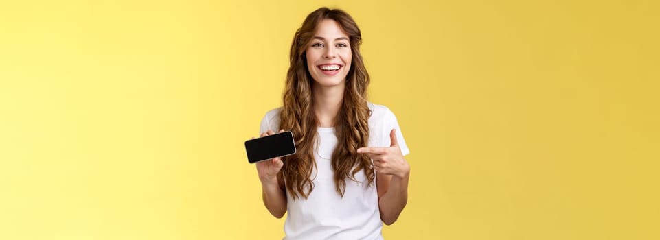 Carefree charismatic cute curly-haired female gamer likes playing smartphone games showing own game score hold mobile phone horizontal pointing display laughing pleased amused. Copy space