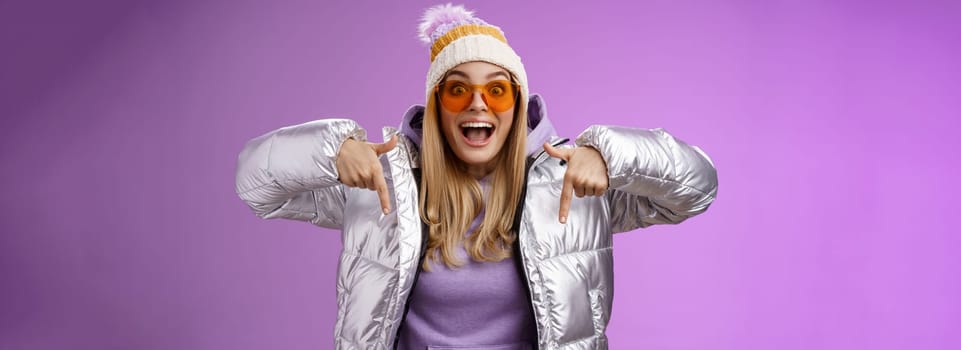 Lifestyle. Excited impressed good-looking blond girl drop jaw amused overwhelmed pointing down index fingers checking out awesome promotion standing surprised thrilled wearing silver winter jacket hat.