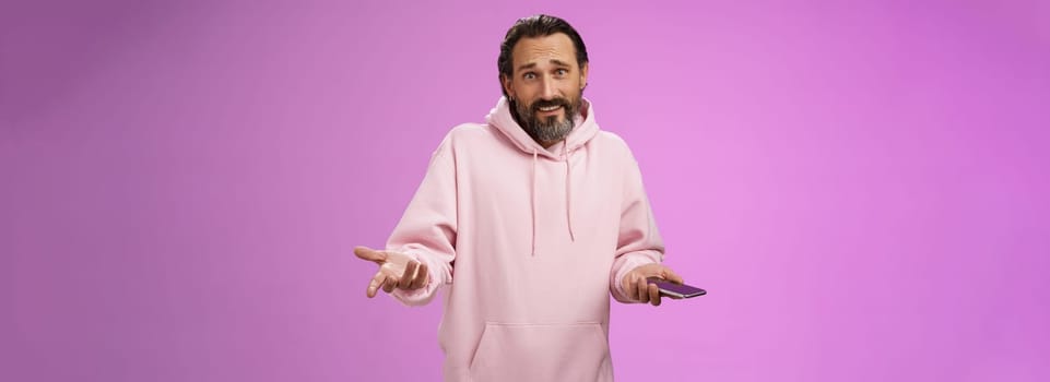 So what. Confused questioned adult bearded stylish guy in pink hoodie shrugging hands spread sideways dismay holding smartphone look clueless unaware camera not know how deal annoying phone calls.