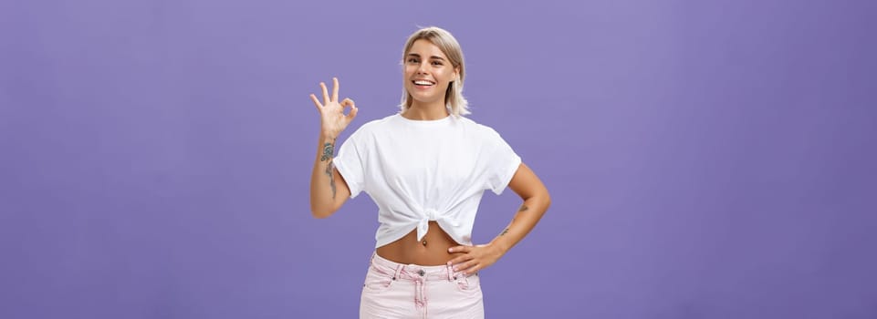 Excellent I like it. Portrait of satisfied good-looking happy girl with fair hair in white t-shirt and shorts showing ok or perfect gesture and smiling broadly holding hand on waist over purple wall.