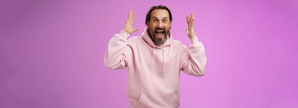 Happy excited lucky adult bearded man celebrating excellent news raising hands gesturing thrilled smiling broaldy triumphing victory win, yelling gladly achieve goal, standing purple background.