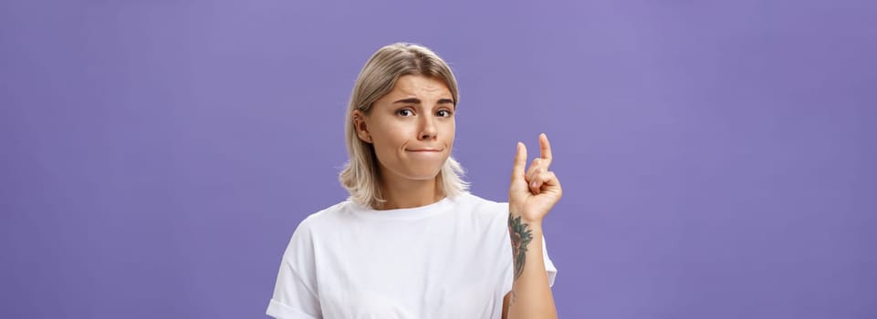 Girl having tiny problem. Concerned attractive blonde girl with tattoo on arm pursing lips in troubled look shaping small or little object, dissatisfied with regret in eyes over purple wall. Copy space