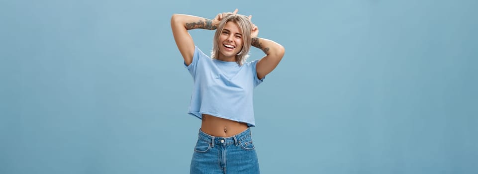 Lifestyle. Studio shot of cute attractive and joyful stylish blond woman with tattoos on arms winking and smiling broadly holding index fingers on head making horns standing in summer outfit over blue wall.