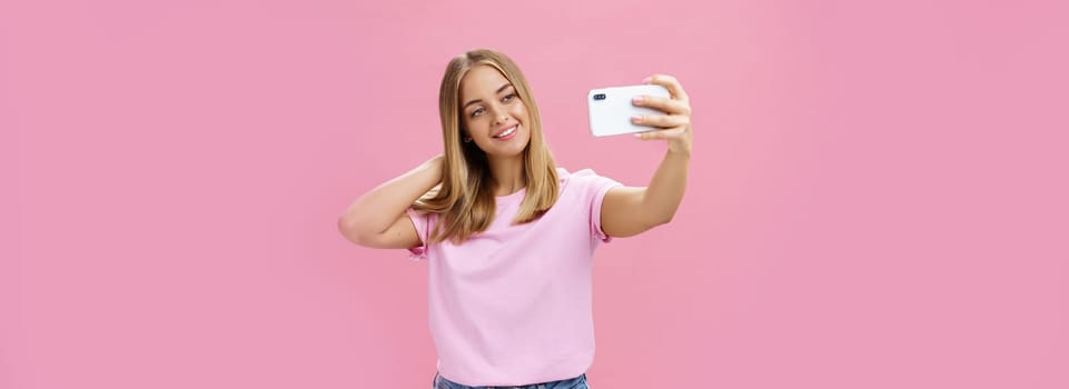 Female beauty blogger taking selfie post new look online. Portrait of charming tanned young woman in t-shirt touching hair gently pulling hand with smartphone near face taking photo over pink wall. Technology concept