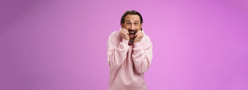 Amused thrilled gasping handsome happy bearded adult man in pink hoodie stooping screaming joyfully receive awesome incredible present standing speechless excited purple background. Copy space