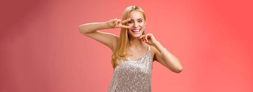 Lifestyle. Charismatic funny amused charming blond girl 25s having fun dancing red background in silver trendy glittering party dress show peace disco gesture near eye smiling laughing carefree.