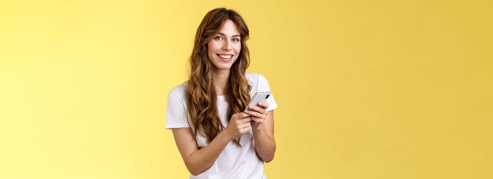 Enthusiastic good-looking urban girl wear white t-shirt standing casual smiling delighted camera texting hold smartphone scroll social media feed stand yellow background browsing network. Lifestyle.