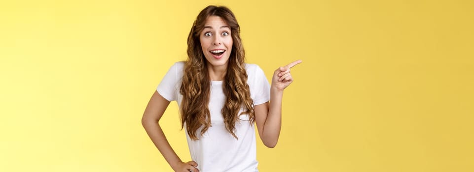 Surprised friendly sociable happy cute girlfriend long curly chestnut haircut smiling impressed gladly pointing left grinning camera toothy astonished expression discuss awesome performance.