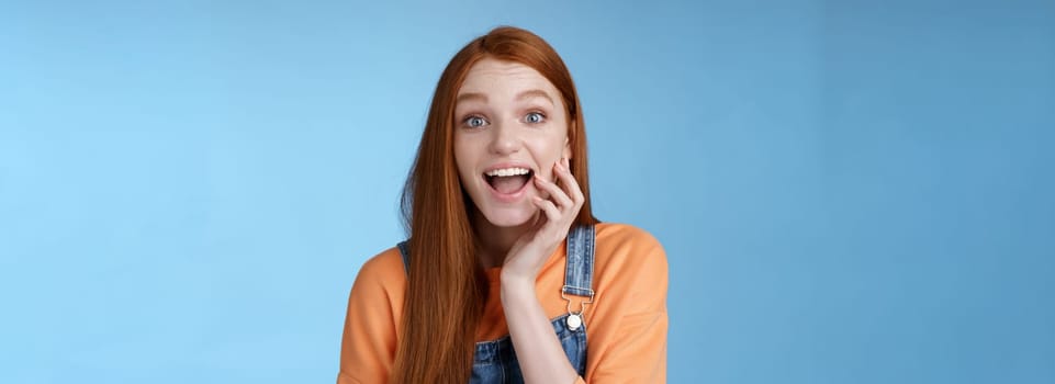 Excited enthusiasitc charismatic redhead female friend look surprised amused touch cheek open mouth thrilled discussing future prom rejoicing standing blue background joyful delighted.