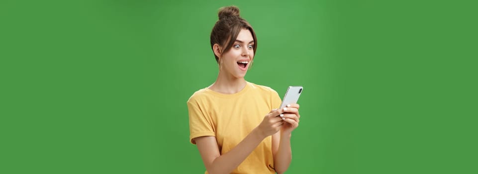 Woman reading surprising satisfying message in smartphone opening mouth from excitement, smiling amazed looking astonished at cellphone screen posing against green background in casual yellow t-shirt. Technology concept