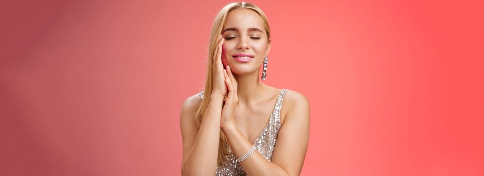 Charming tender lovely coquettish blond woman close eyes smiling relaxed flirty touch face dreaming heartwarming romantic moment, standing red background silver evening dress seducing.
