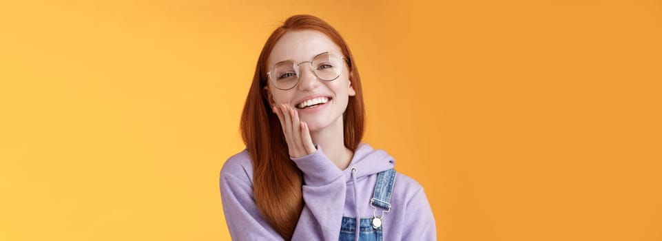 Charismatic happy tender redhead smiling european girl 20s glasses hoodie denim overalls having fun enjoying summer holidays chilly evening laughing joyfully touch pure clear skin, orange background.