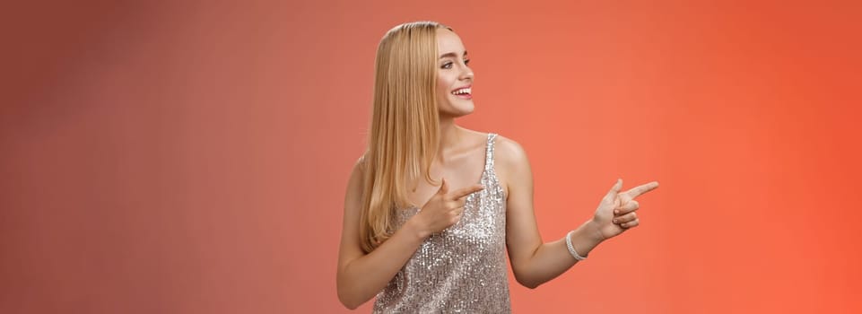 Joyful carefree young wealthy glamour blond woman in glittering silver dress enjoying awesome party dancing turning pointing left smiling broadly waiting girlfriend bring drinks, red background.