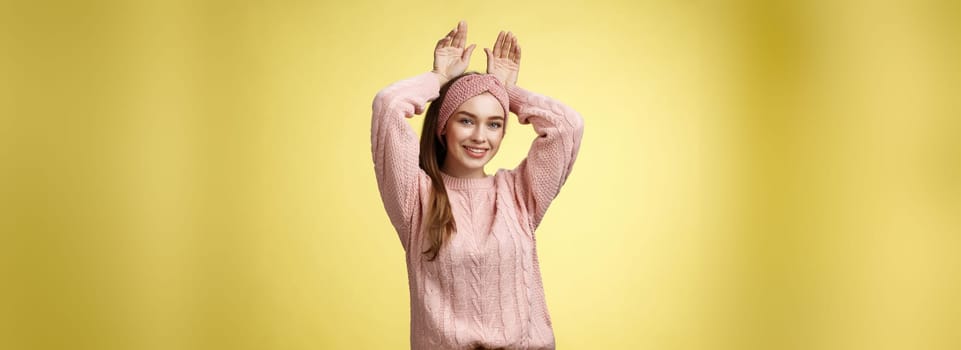 Nice charming playful young european female having fun, playing mimicking bunny showing rabbit ears with hands behind head, smiling cute, tender, happy greeting boyfriend welcome back ove yellow wall.