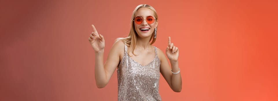 Stylish carefree fabulous young blond woman in silver dress red sunglasses having fun dancing joyfully dance-floor nightclub smiling broadly enjoying spend time friends, standing studio background.