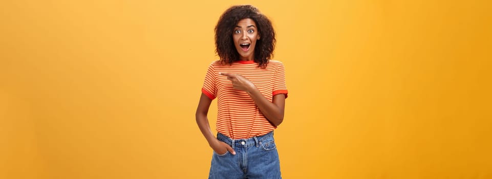 Hey awesme copy space there. Portrait of impressed and surprised enthusiastic young african american female with afro hairstyle pointing right astonished looking energized and curious over orange wall.