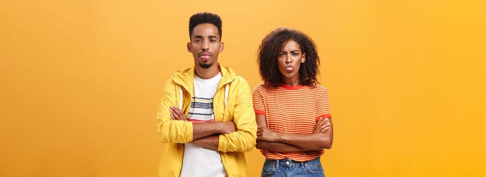 Indoor shot of african american siblings being displeased and annoyed showing bad tempber behaving childish sticking out tongue standing together with crossed hands on chest over orange background. Lifestyle.