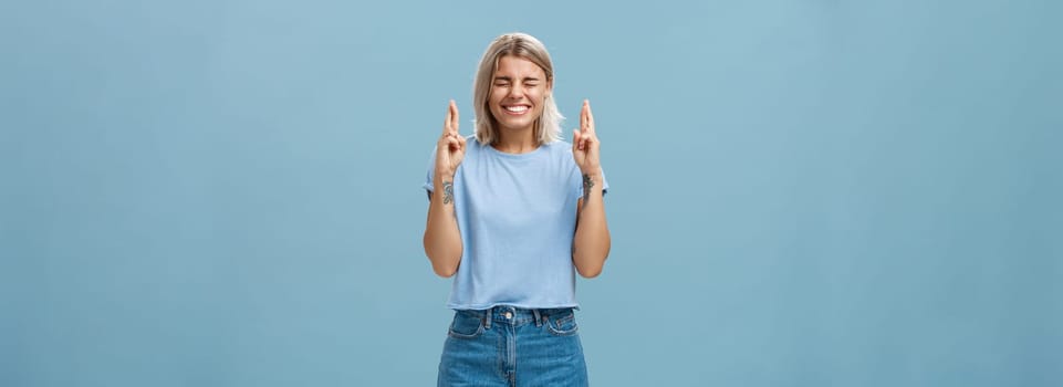Optimistic faithful good-looking young woman with blond hair and tattoos smiling joyfully crossing fingers for good luck waiting for dream come true making wish while standing over blue background. Copy space