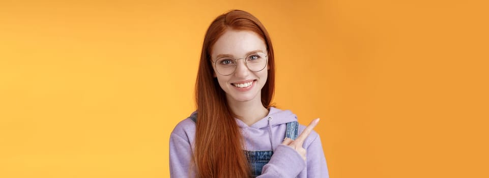 Friendly good-looking modern redhead young girl pointing left index finger showing awesome place suggesting go hang out smiling joyfully casually talking discuss new product, orange background.
