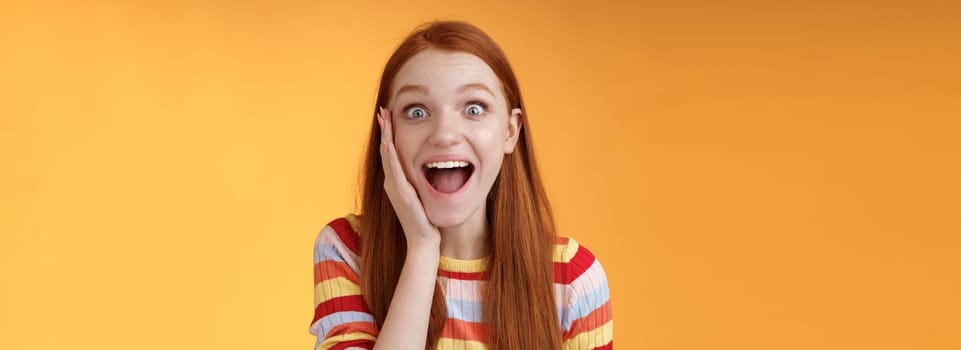 Amused happy smiling good-looking redhead woman screaming happily touch cheek astonished receive good perfect news triumphing feeling excited look thrilled camera, standing orange background.