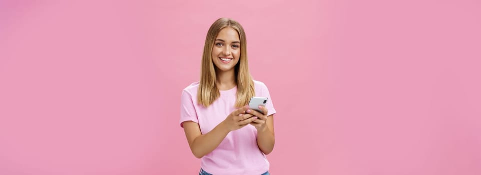 Woman calling taxy via smartphone asking friend address smiling cheerfully at camera holding phone with both hands over chest getting in touch with clients via messages standing over pink wall. Technology concept