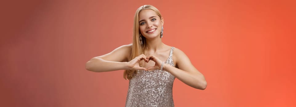 Romantic passionate charming lovely blond glamour woman in silver dress brilliants show heart love gesture express sympathy passion, adore relationship smiling delighted, standing red background.