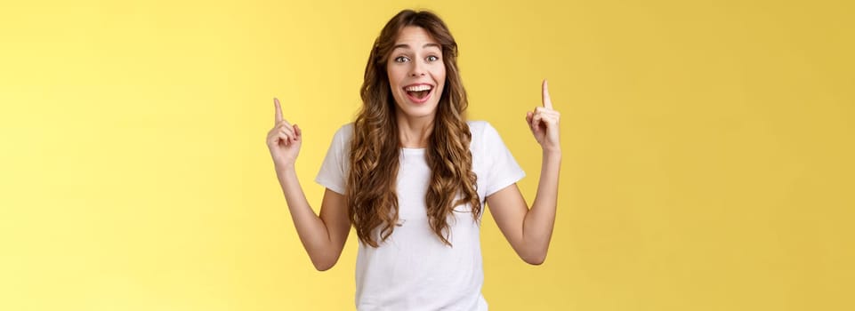 Surprised cheerful celebrating girl lively discuss near awesome event pointing up index fingers smiling broadly fascinated look temtation admire great concert stand yellow background impressed.