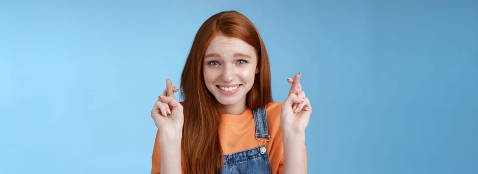 Lifestyle. Silly young pretty redhead girl blue eyes freckles making wish cross fingers good luck smiling hopeful eager get best results waiting desires fulfill miracle happen anticipating blue background.