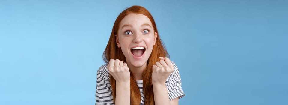 Yes awesome news great. Attractive surprised redhead girl cheering happy clench fists yelling yeah accomplish goal wide eyes excited celebrating win lottery triumphing joyfully, blue background.