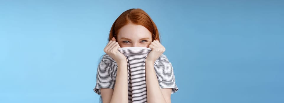 Silly flirty amused attractive playful redhead girlfriend hiding face pulling t-shirt head squinting devious mysteriously giggle laughing hope disguise pranking friend standing blue background.