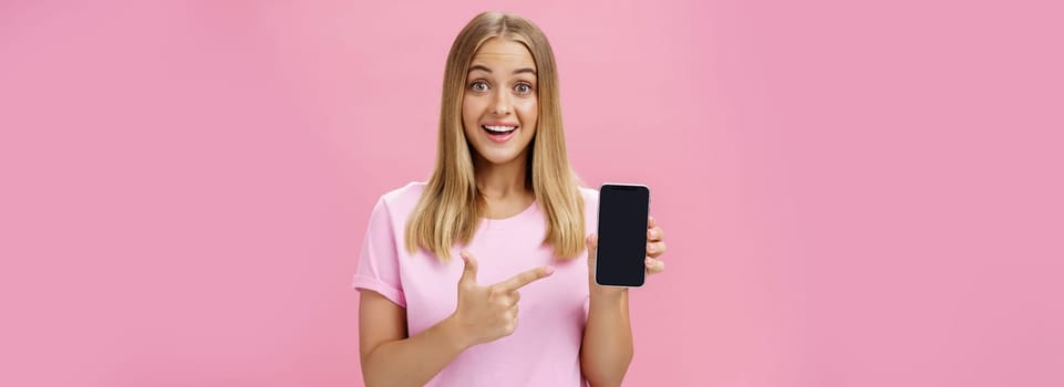Cheerful attractive and pleasant woman promoting cool app or smartphone holding cellphone and pointing at device screen smiling amused and impressed standing over pink background. Technology concept